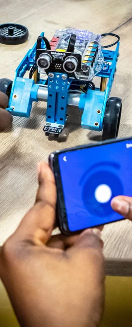 Student using a smartphone to control a small blue robotic vehicle, testing its movements on a table in a computing lab. Student using a smartphone to control a small blue robotic vehicle, testing its movements on a table in a computing lab.